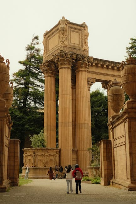 The image depicts a historic outdoor setting with large, ornate stone columns supporting detailed carved capitals and a high, rectangular structure atop the columns, resembling ancient ruins. Surround